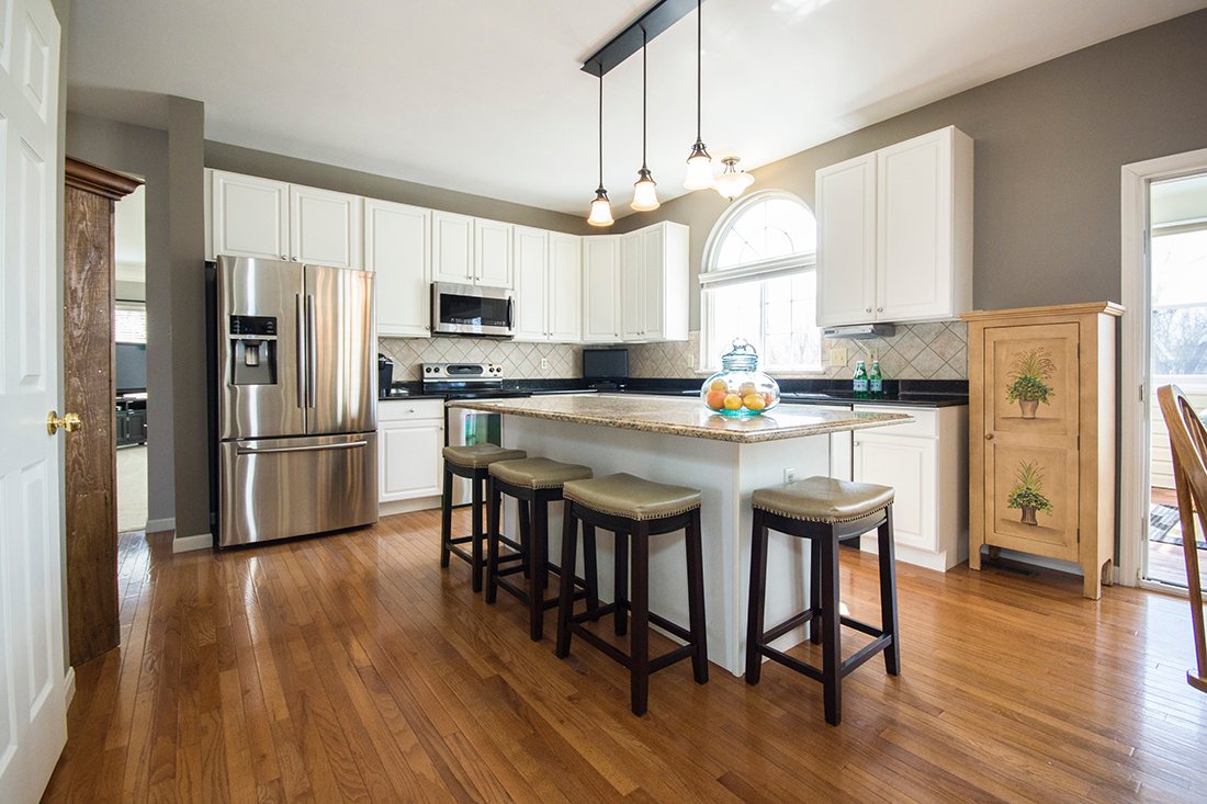 A white kitchen with a large island and three bar stools, pendant lights above the island and an American fridge-freezer