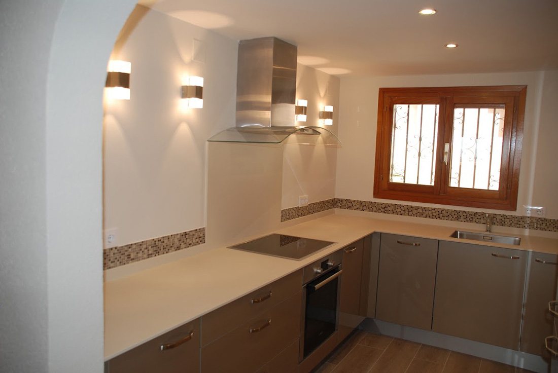 A newly fitted kitchen in taupe and white with a stainless steel chimney extractor, an induction hob and a shuttered window