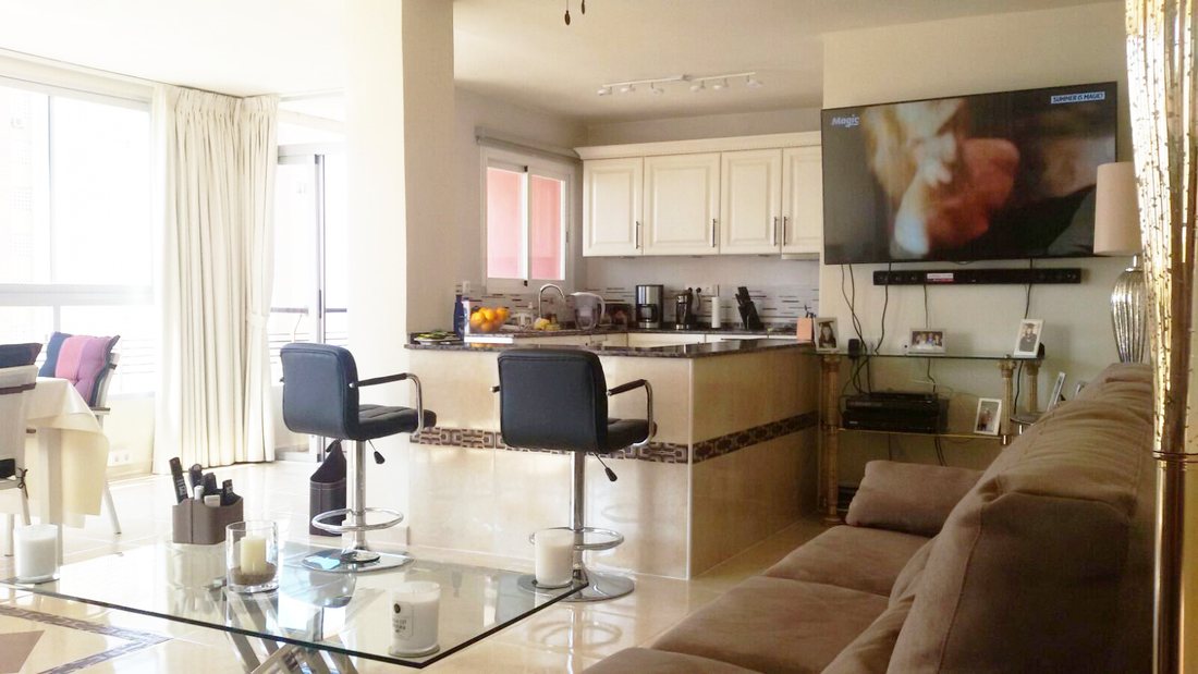 An apartment living room looking through to the kitchen, with a breakfast bar, dark stools and balcony doors beyond