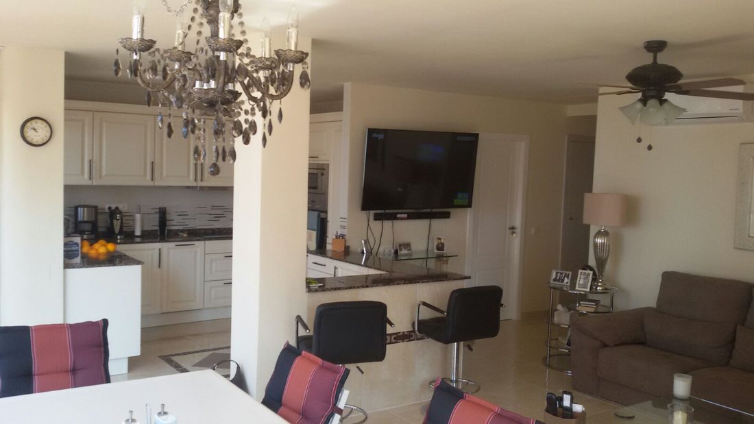 An open-plan kitchen and living room with white units, a breakfast bar with dark stools, a chandelier and a wall-mounted television