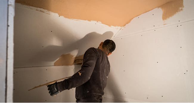 A plasterer in dark clothing smoothing a freshly boarded wall with a trowel, plaster patches visible on the plasterboard
