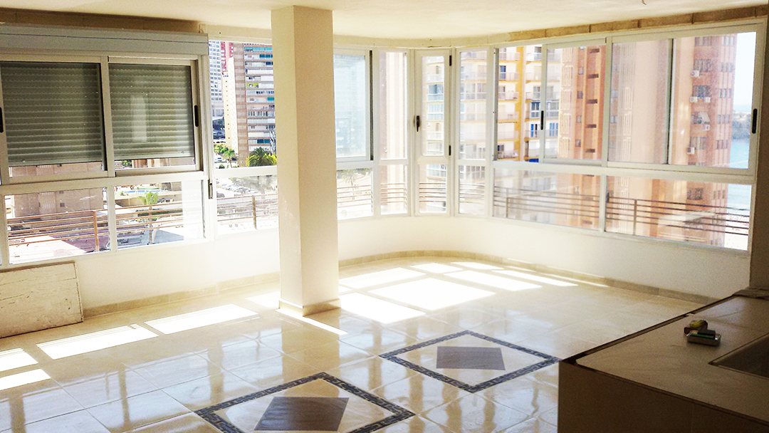 A bright tiled apartment room with a full wall of white-framed windows and roller blinds above the city