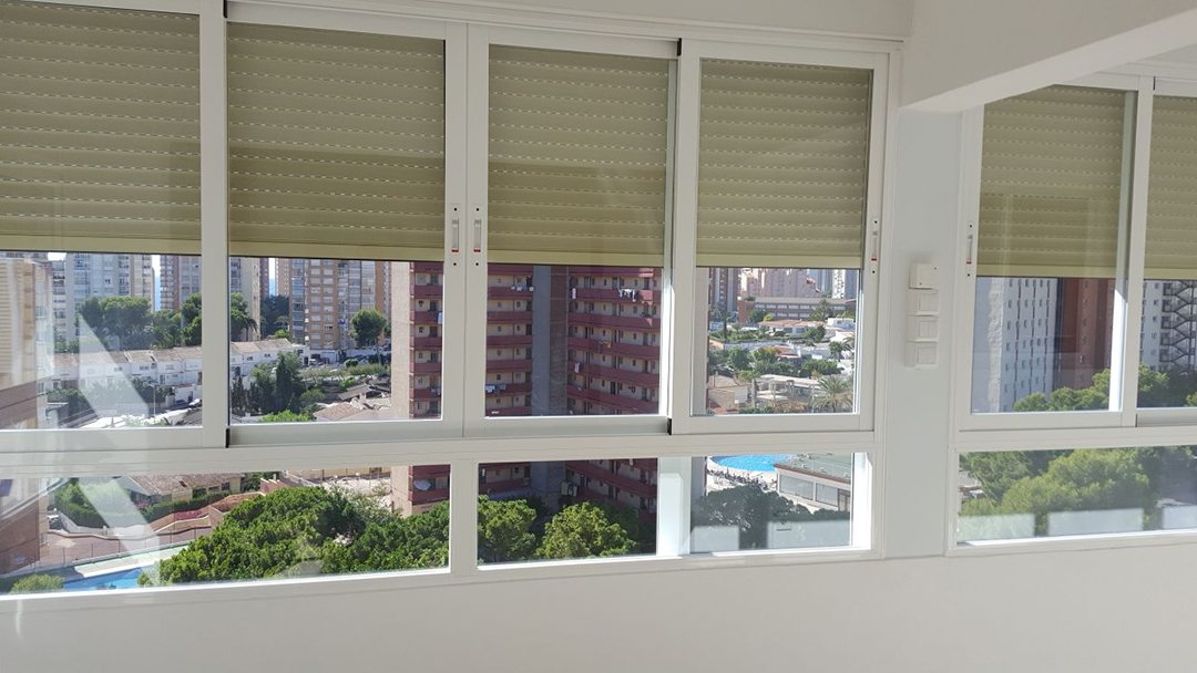 A row of white-framed windows with beige roller blinds overlooking trees and tower blocks